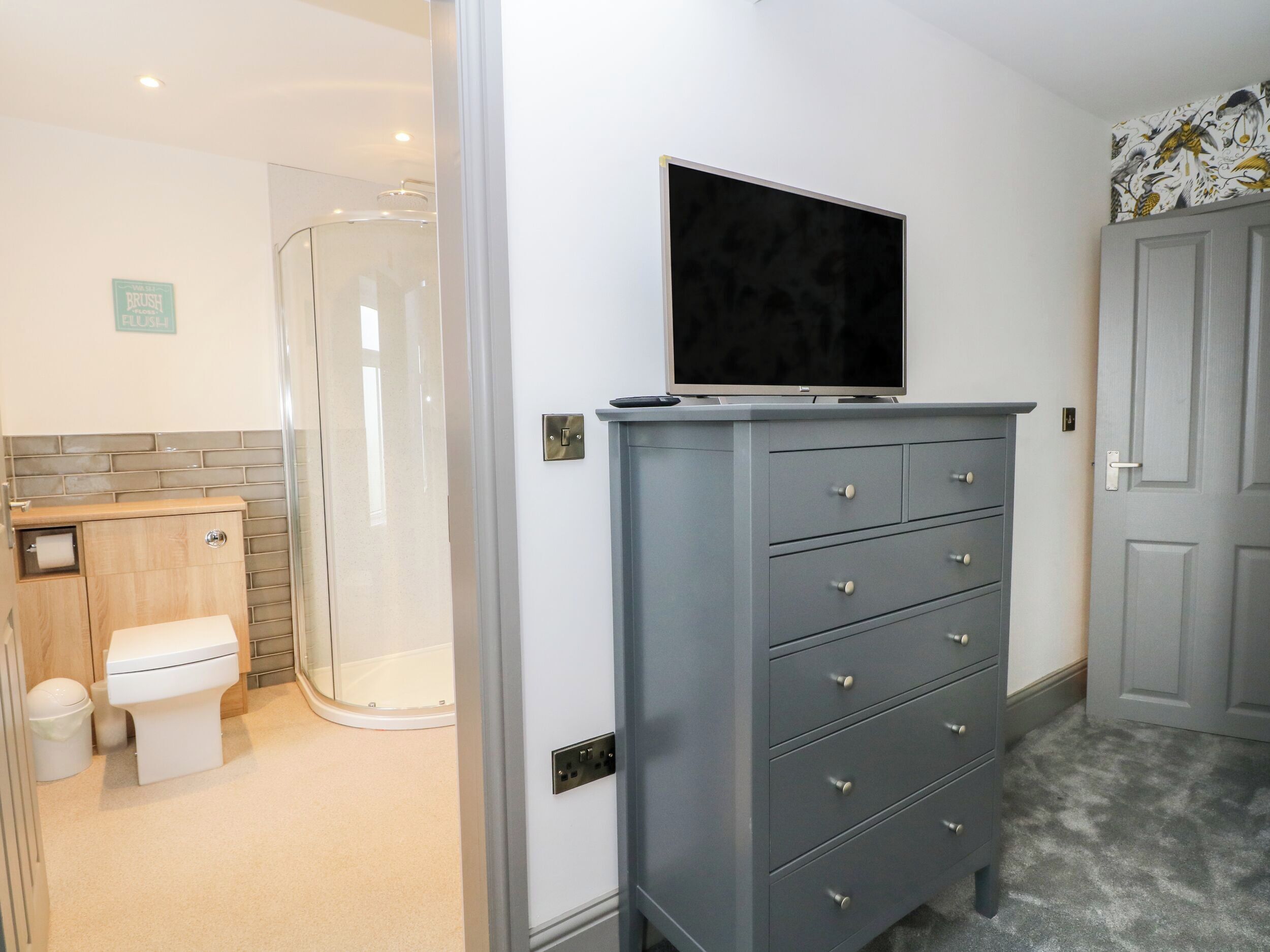 A bathroom with a shower, toilet, vanity, and a grey dresser with a flat-screen TV mounted above it.