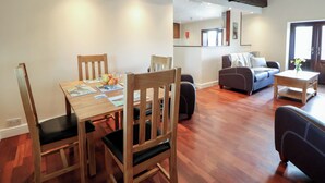 Uma sala de jantar com mesa e cadeiras de madeira, uma cozinha ao fundo e uma sala de estar com sofá e uma mesinha.