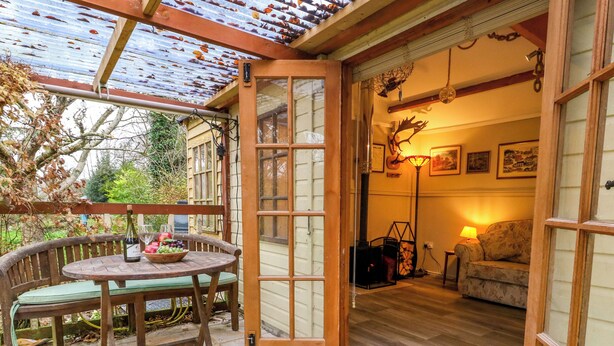 A wooden table with a bottle and fruit, a bench, and a wooden door leading to a room with a sofa and lamp. - The Cabin at Little Timbers (Ringwood)