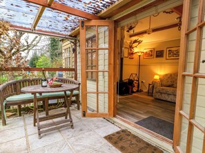 A wooden table with a bottle and fruit, a bench, and a wooden door leading to a room with a sofa and lamp. - The Cabin at Little Timbers (Ringwood)