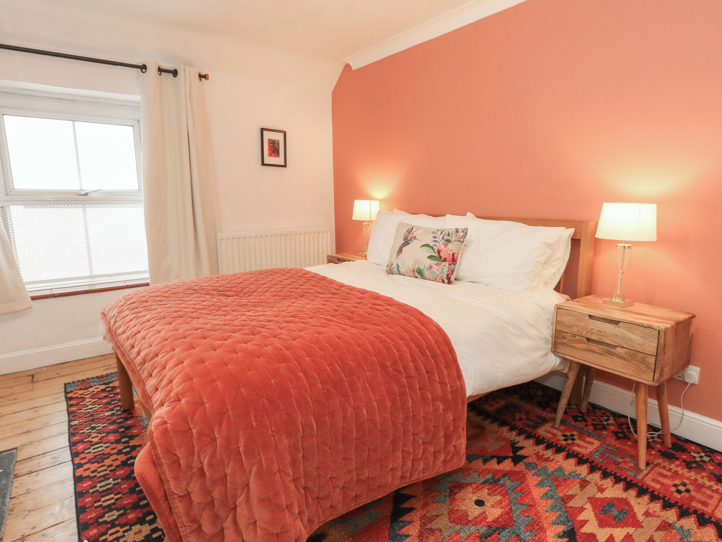 A bedroom with a large bed, two bedside tables with lamps, a window with curtains, and a patterned rug.