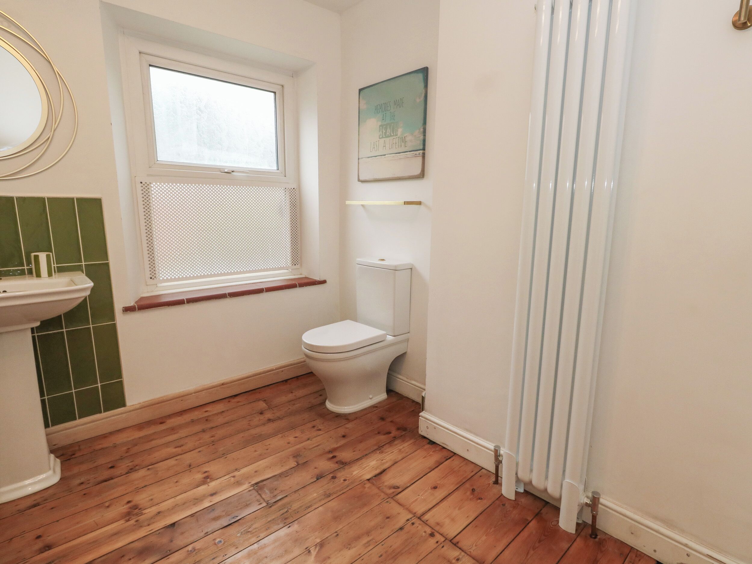 A bathroom with a wooden floor, a white toilet, a window with a white blind, and a green-tiled wall.
