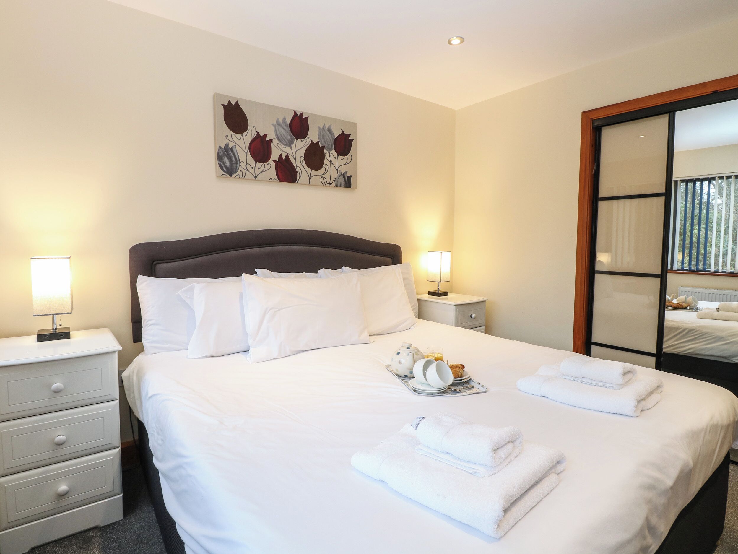 A neatly made bed with white linens, a bedside table with a lamp, a nightstand with a lamp, and a painting on the wall.