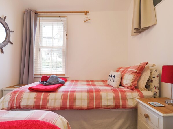 A bedroom with two beds, a window with blinds, a bedside table with a lamp, and a wall clock. - Puffins (Sidmouth)