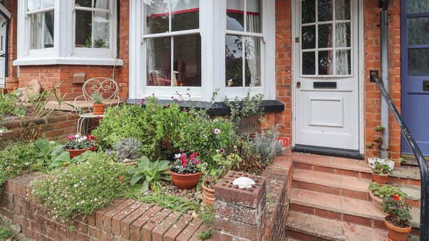 A brick house with a white door, a bay window with curtains, and a small garden with potted plants. - Puffins (Sidmouth)