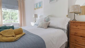 A neatly made bed with a blue and white patterned bedspread, a wooden nightstand with a lamp, and a dresser with a mirror. - Puffins (Sidmouth)