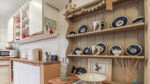 A kitchen with wooden cabinets, a microwave, a toaster, and a coffee maker. There are shelves with plates and bowls. - Puffins (Sidmouth)