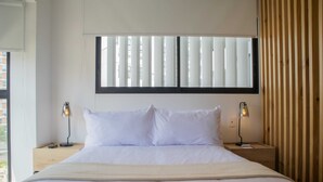Una cama bien hecha con sábanas blancas, dos lámparas de noche, una ventana con persianas y un panel de pared de listones de madera.
