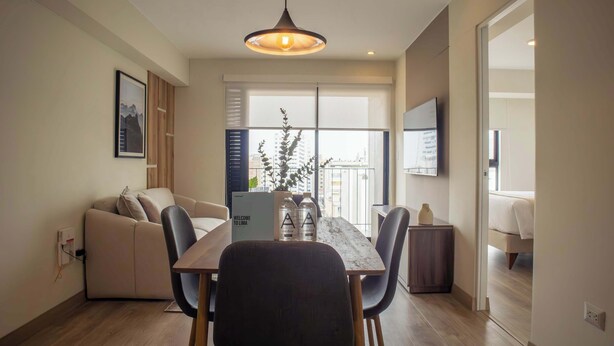 A modern living room with a sofa, a dining table with chairs, a TV mounted on the wall, and a large window with blinds.