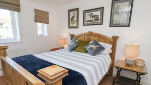 A neatly made bed with a striped bedspread and pillows, a wooden headboard, a bedside table with a lamp, and framed wall art above the bed. - Coastal Retreat (Seahouses)