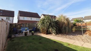 A residential backyard with a grassy lawn, a wooden fence, a table with chairs, and a large plant. - Coastal Retreat (Seahouses)