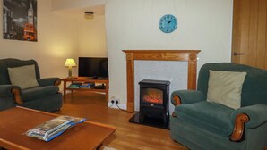 Ein Wohnzimmer mit zwei grünen Sesseln, einem Holztisch, einem Kamin, einem Fernseher und einer Wanduhr.