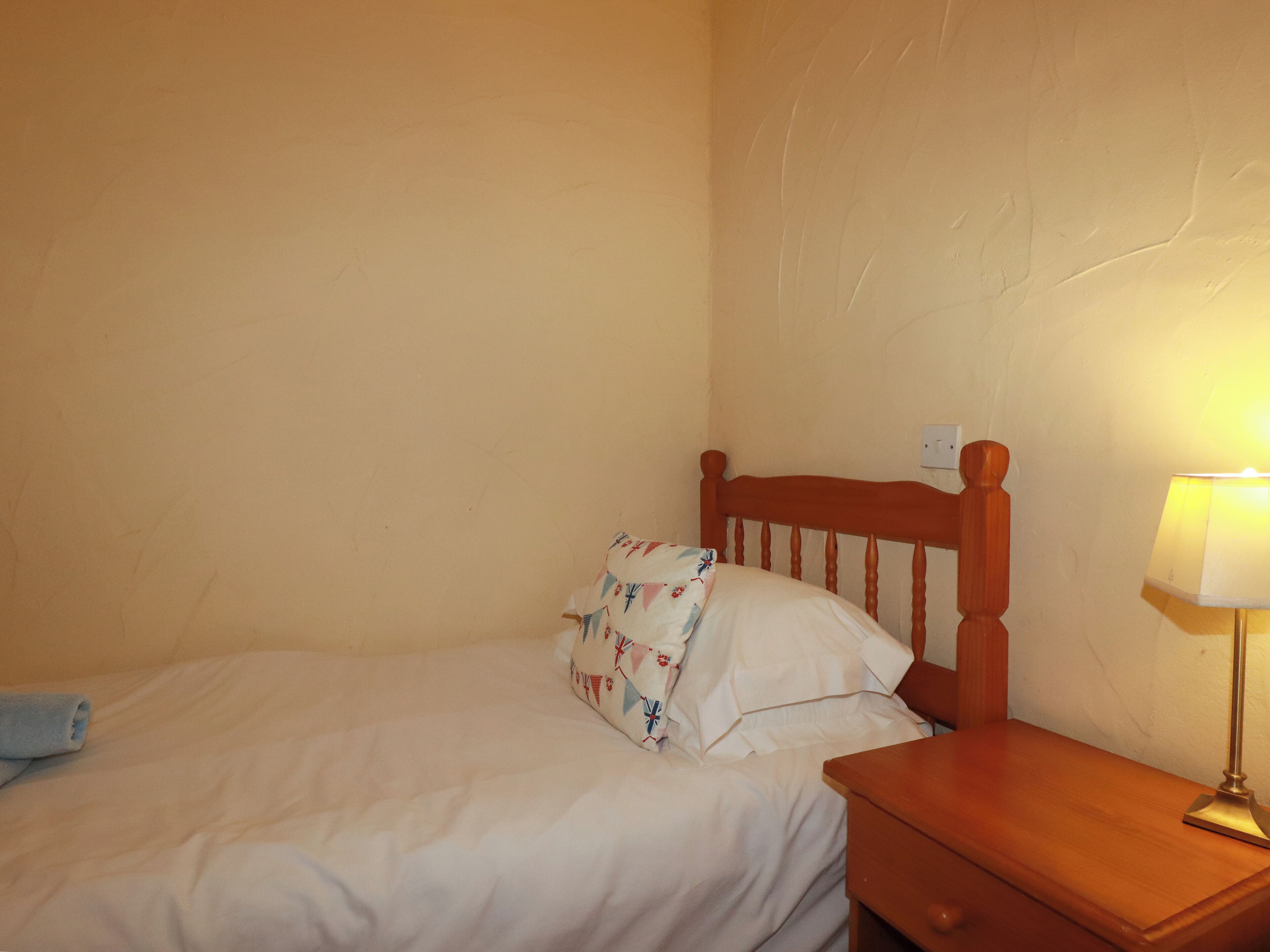 Ein Einzelbett mit Holz-Kopfstück, ein Nachttisch mit Lampe und eine Steckdose an der Wand.