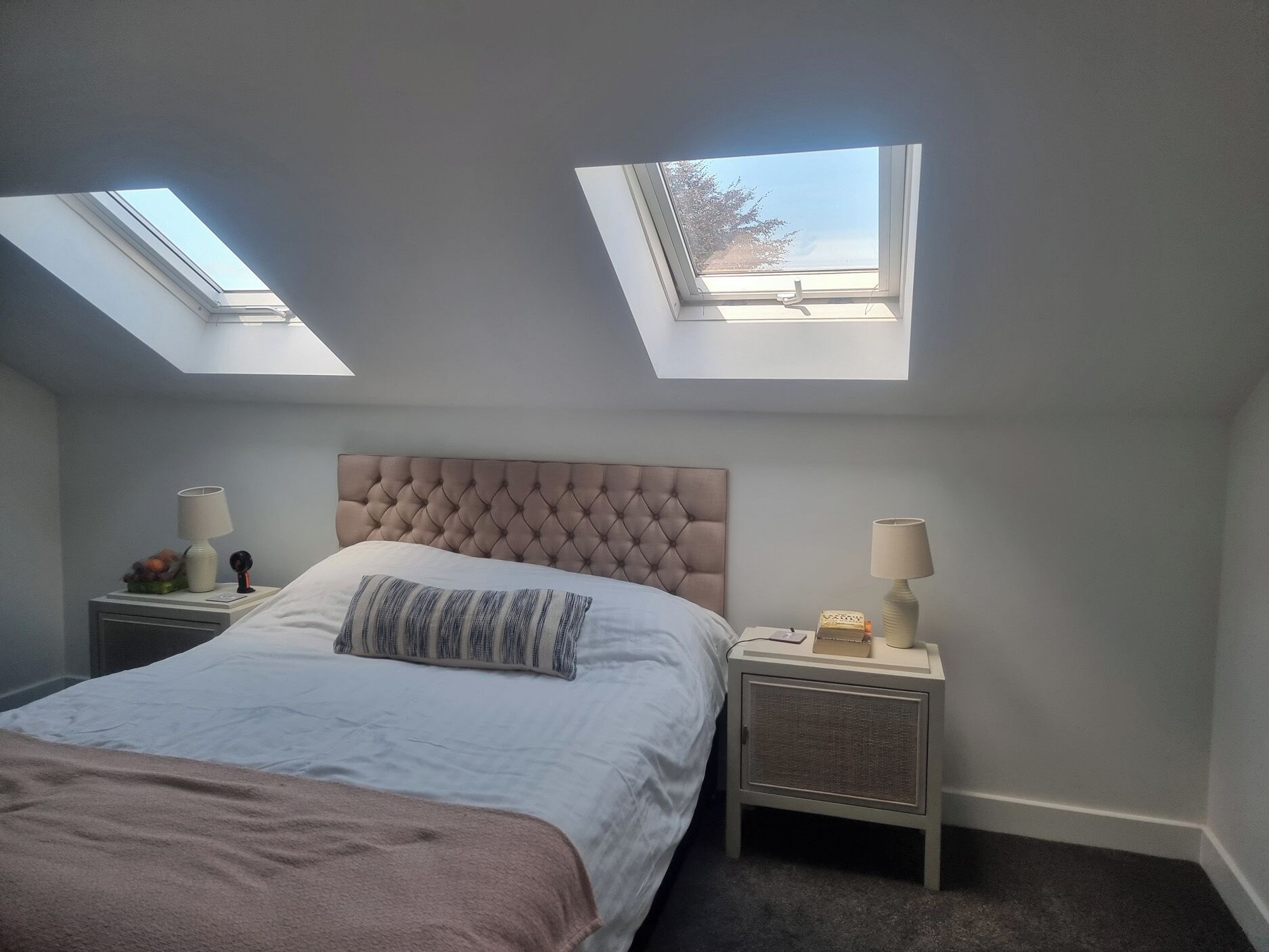 Ein Schlafzimmer mit einem Bett, zwei Nachttischen, einem gepolsterten Kopfteil und Dachfenstern.