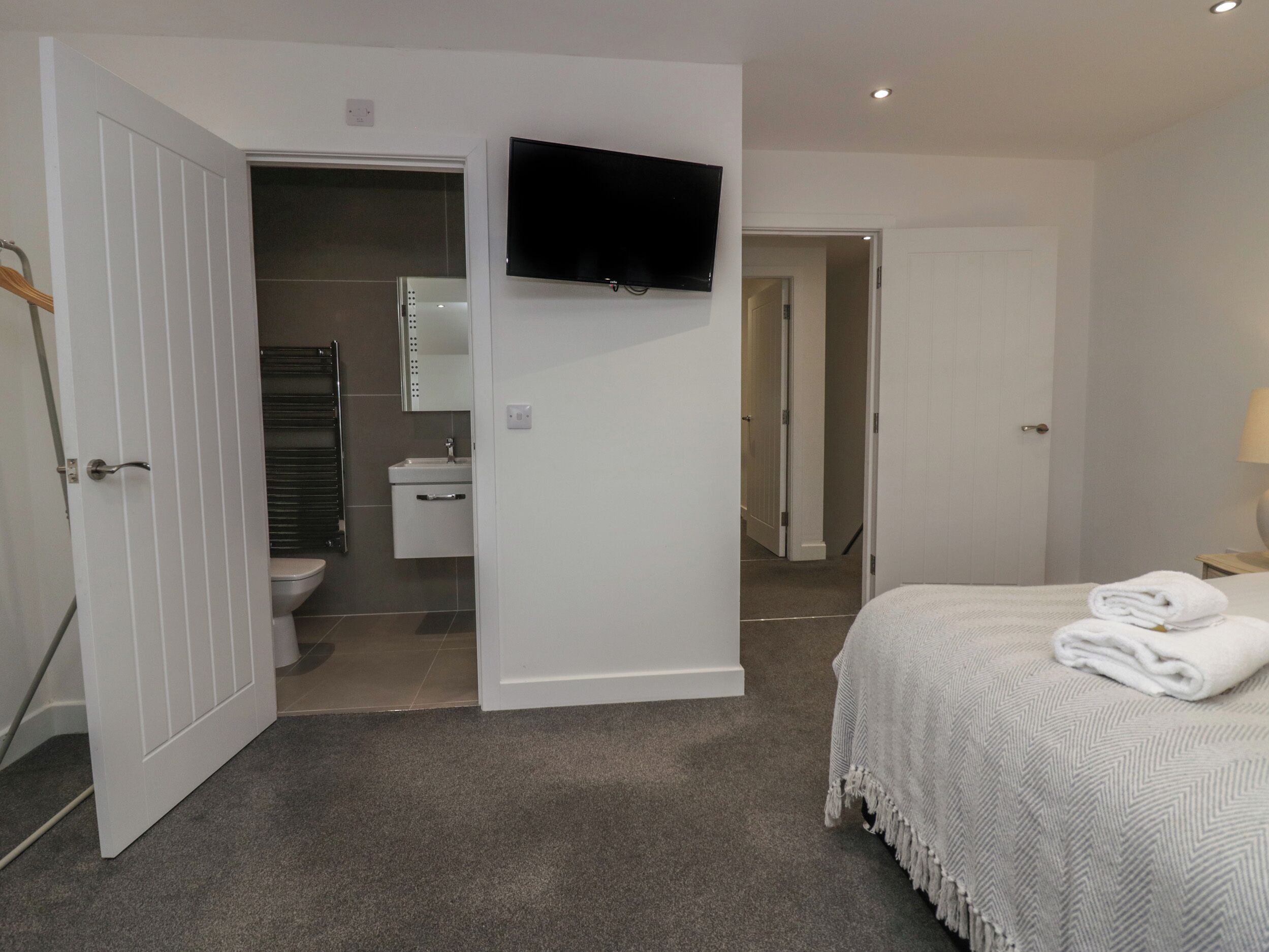 A bedroom with a bed, a TV mounted on the wall, a bathroom with a toilet and sink, and a door leading to another room.