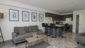 A modern living room with a grey sofa, a wooden coffee table with wine glasses, and a kitchen in the background. - Edgemere Retreat (Grange-over-Sands)