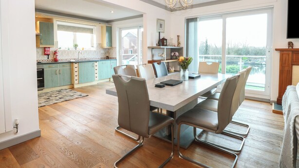 A modern kitchen with teal cabinets, a dining area with a large table and chairs, and a view of a body of water through large windows. - White Gables (Norwich)