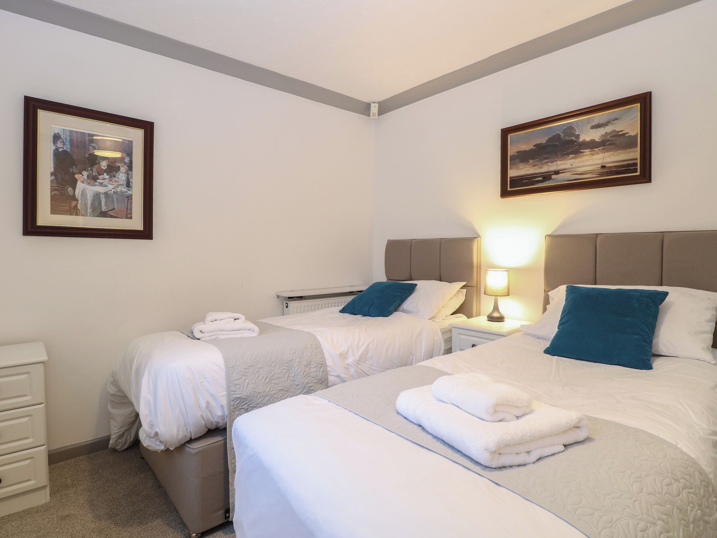 A hotel room with two beds, white bedding, a nightstand with a lamp, a framed picture on the wall, and a small table with a folded towel.