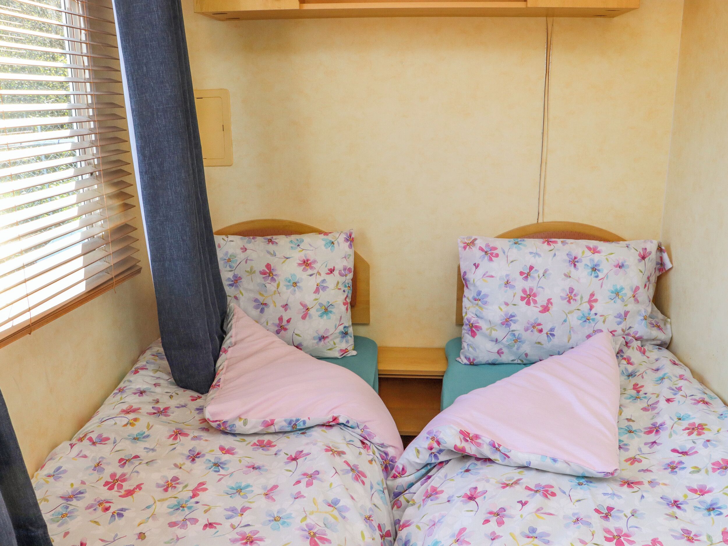 A small, compact room with two single beds, each with floral-patterned bedding and pillows. There is a window with blinds on the left side, and a wooden shelf above the beds.