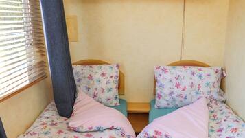 A small, compact room with two single beds, each with floral-patterned bedding and pillows. There is a window with blinds on the left side, and a wooden shelf above the beds.