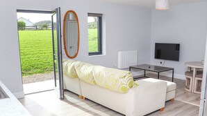 En moderne stue med sofa, TV, spisebord og udsigt til et grønt felt gennem døren.