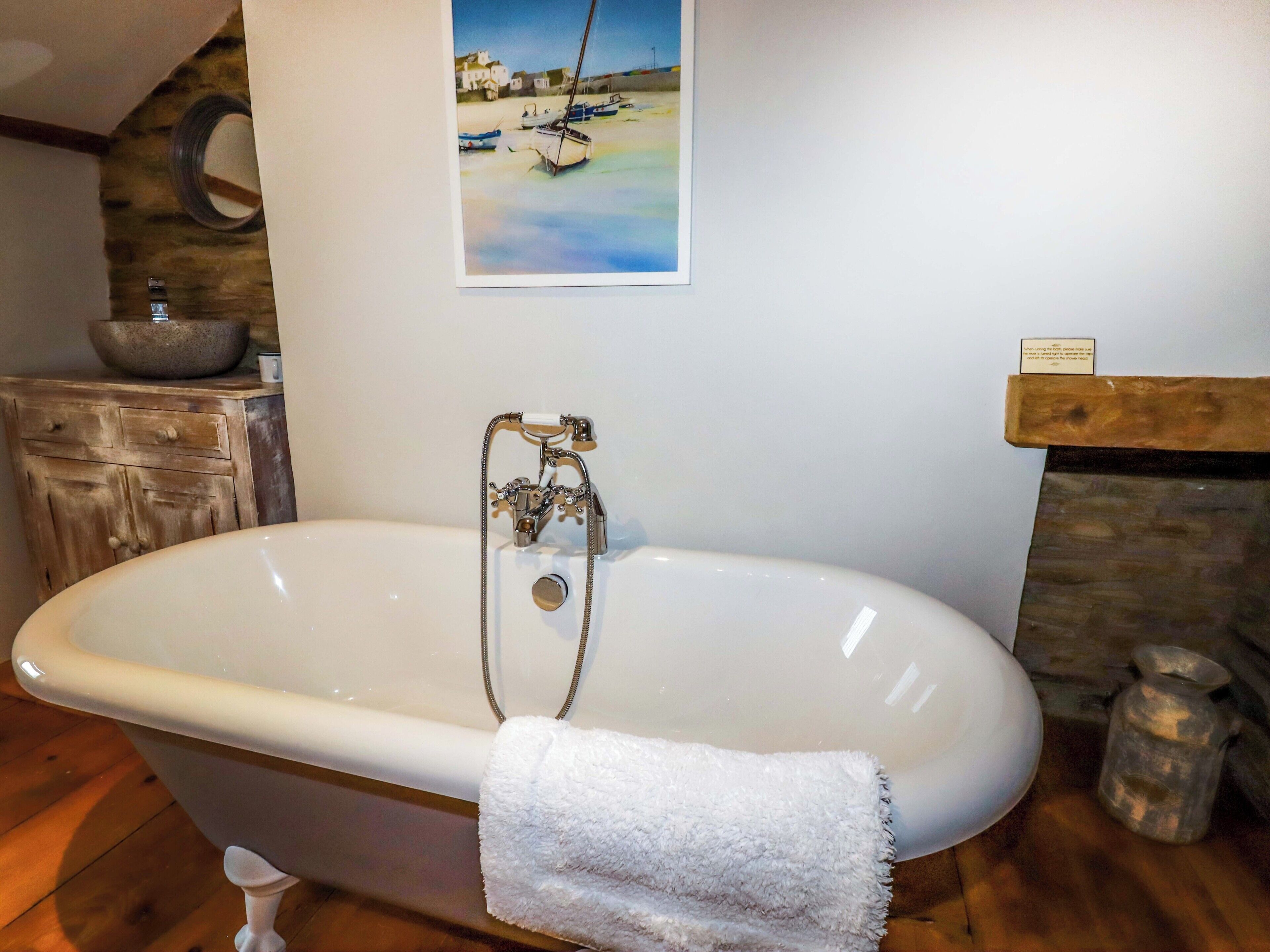 A bathroom with a freestanding bathtub, a wooden cabinet, a stone wall, and a painting of boats on the wall.