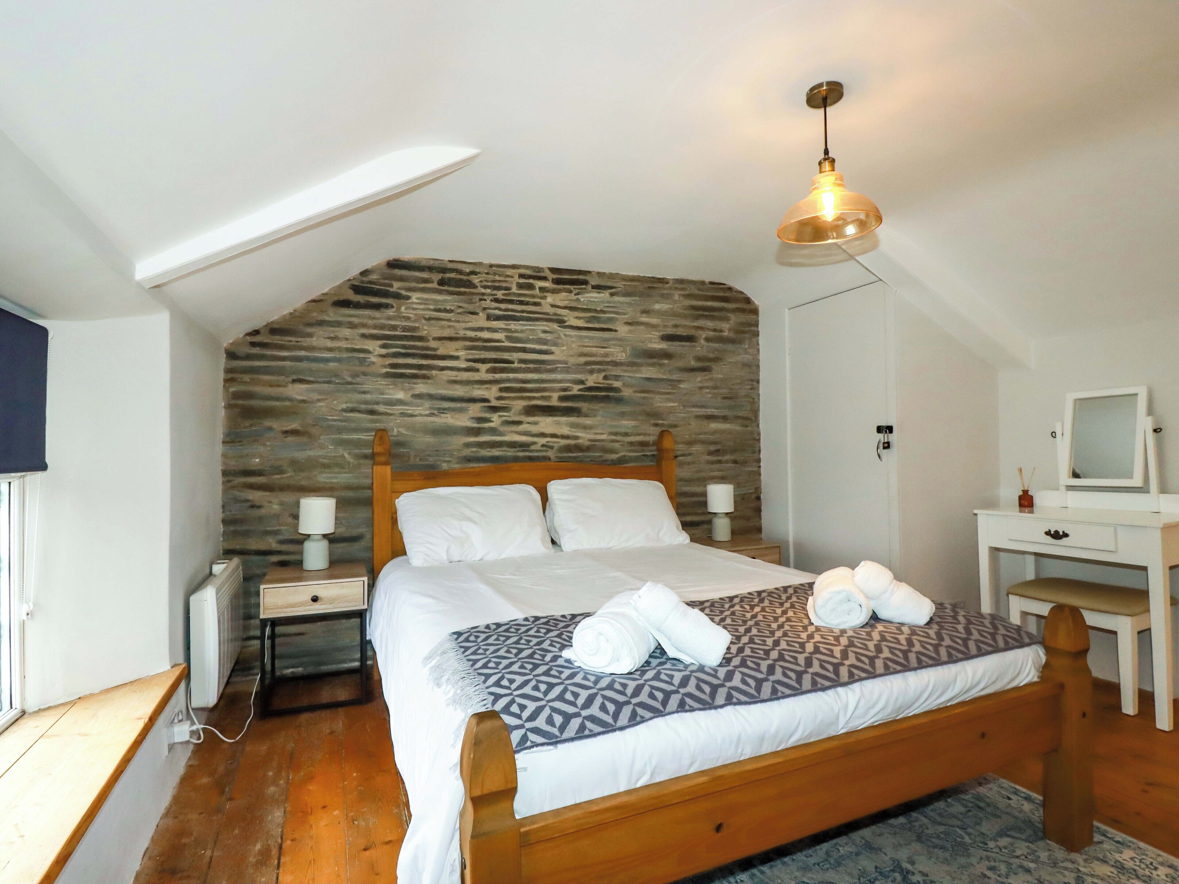 Een slaapkamer met een schuine nok, een stenen muur, een houten bed, een nachtkastje met een lamp, een bureau met een spiegel en een radiator.