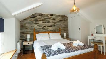 Een slaapkamer met een schuine nok, een stenen muur, een houten bed, een nachtkastje met een lamp, een bureau met een spiegel en een radiator.