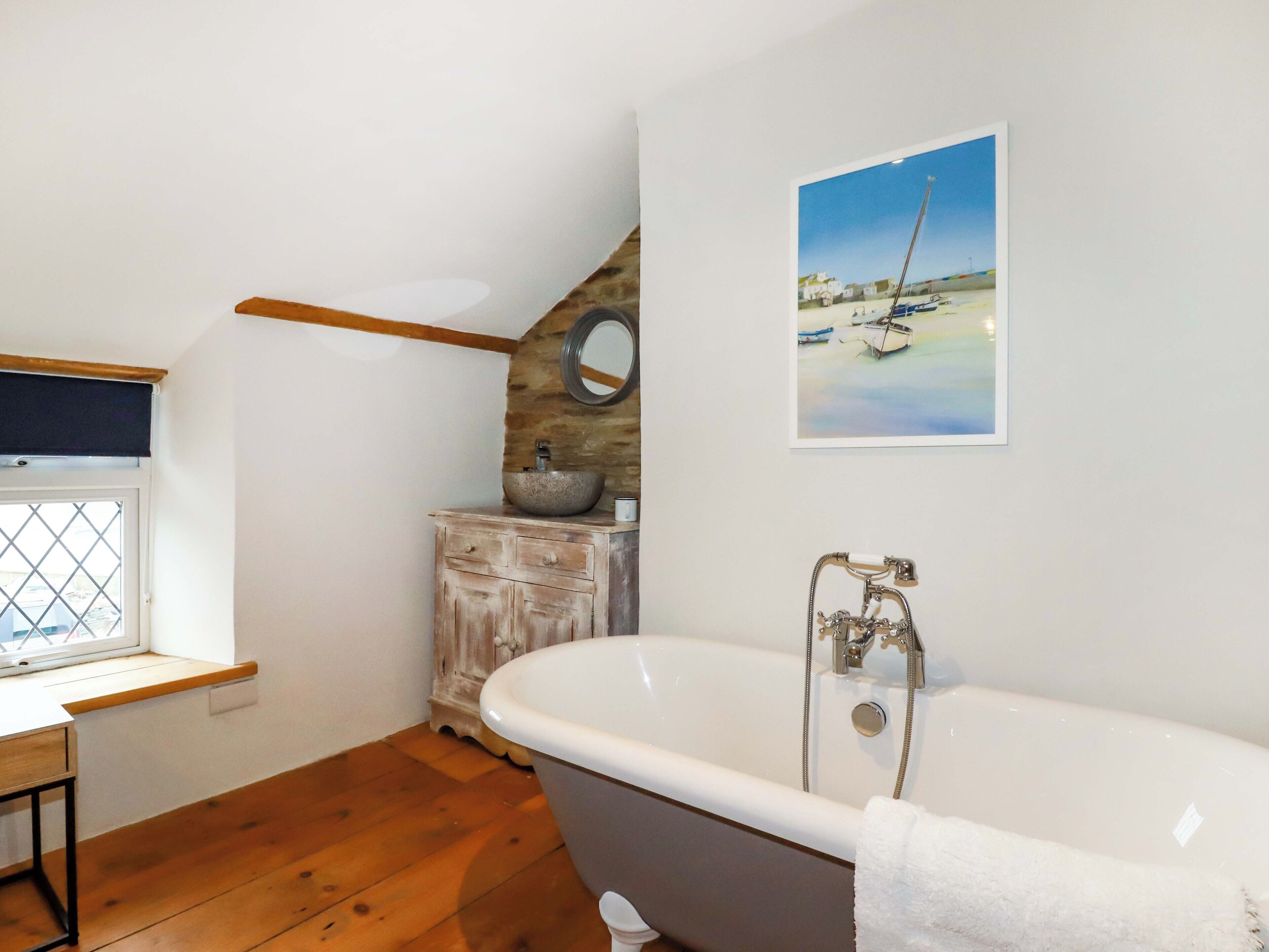A bathroom with a freestanding bathtub, a wooden vanity, a window with a view, and a painting of boats on the wall.