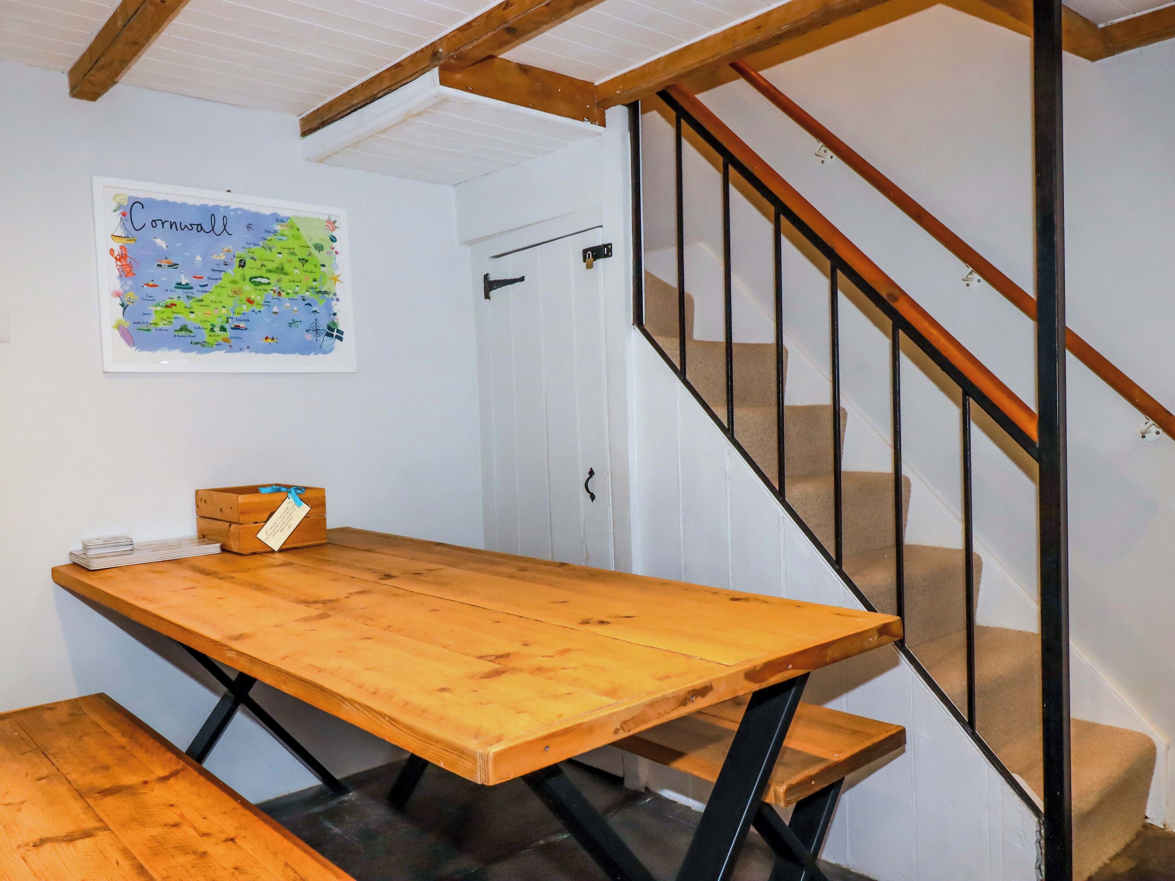 Ein Holztisch, an der Wand eine Karte von Cornwall, eine Treppe mit schwarzen Geländern und eine Tür.