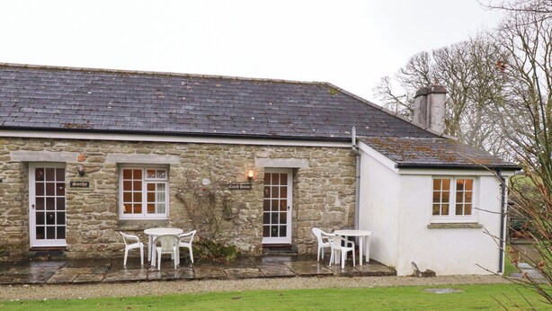 A stone building with two windows and a door, a sign reading "Smith", and a small outdoor seating area with white chairs and a table. - The Coach House (Camelford)