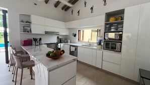 Eine moderne Küche mit weißen Schränken, einer Marmorarbeitsplatte und einer zentralen Kochinsel mit Obst. Es gibt Korbstühle, eine Kaffeemaschine und eine Mikrowelle.