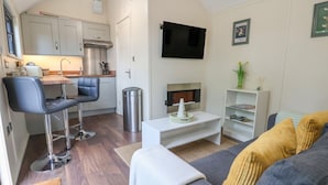 Un salon moderne avec un canapé gris, une table basse, un téléviseur fixé au mur et un coin cuisine équipé de placards et d’une cuisinière.