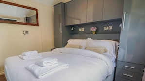 Un lit bien fait, avec du linge de lit et des serviettes blanches, une armoire grise à deux tiroirs et un petit vase avec des fleurs accroché au mur.