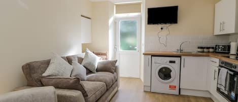 A compact living space with a sofa, a washing machine, a kitchen area, and a TV mounted on the wall.