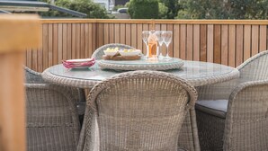 A round glass table with wicker chairs, a bottle of wine, and glasses on a wooden deck with a view of houses and trees. - Hoheck (Pwllheli)