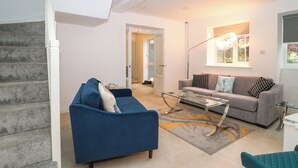 A modern living room with a blue sofa, a grey sofa, and a glass coffee table. There is a staircase on the left and a door leading to another room. - 4 Noel Court (Chipping Campden)