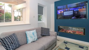 A modern living room with a grey sofa, patterned cushions, a TV displaying a news program, and a glass coffee table. - 4 Noel Court (Chipping Campden)