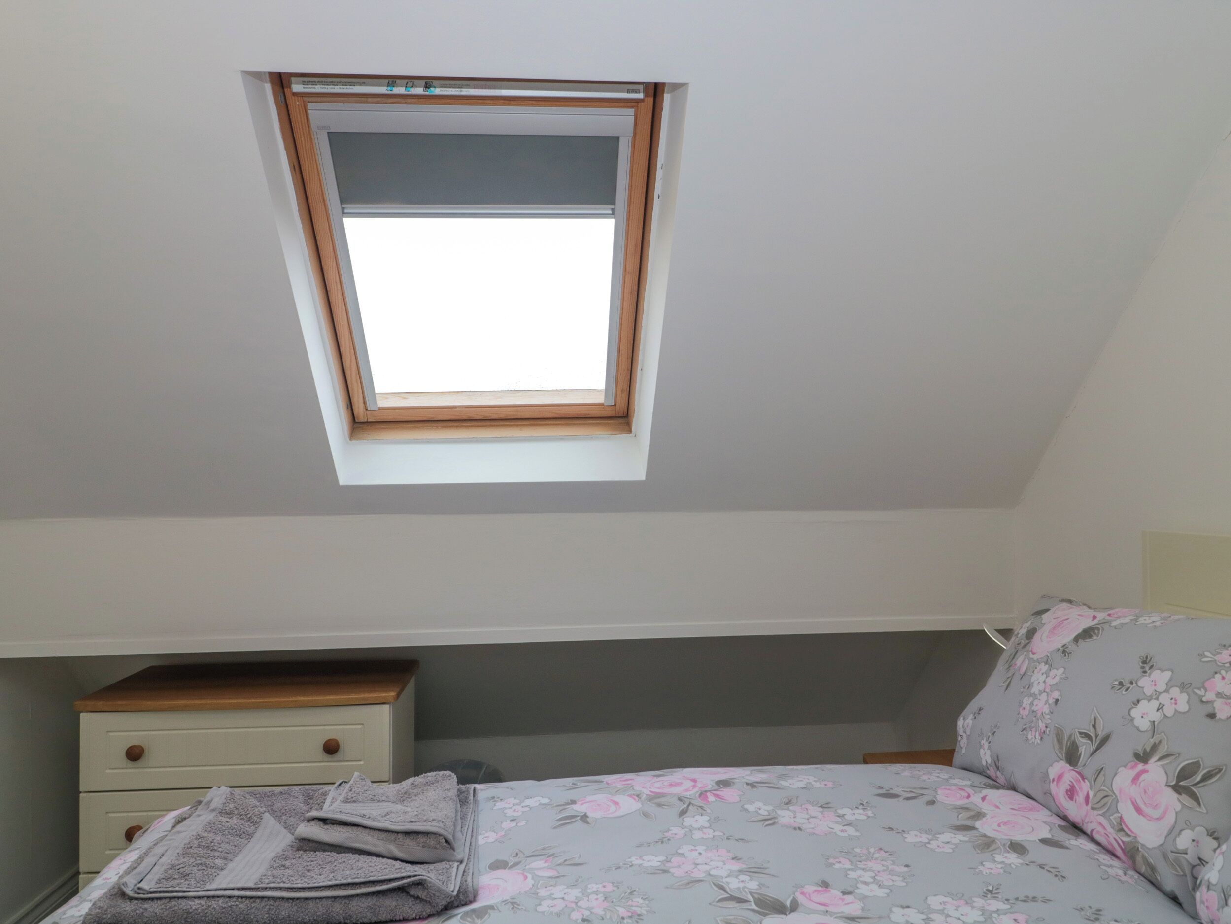 Ein Schlafzimmer mit Dachfenster, einem Bett mit floralem Bettzeug, einem Nachttisch und einer kleinen Kommode.