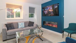 A modern living room with a grey sofa, a glass coffee table, a blue accent wall, a fireplace, and a television mounted on the wall. - 4 Noel Court (Chipping Campden)