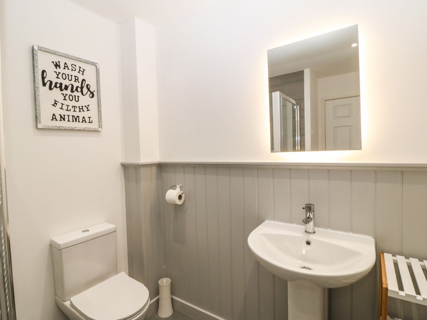 Un baño con inodoro, lavabo y un espejo sobre él. Hay un dispensador de papel higiénico y un rollo de papel en la pared.