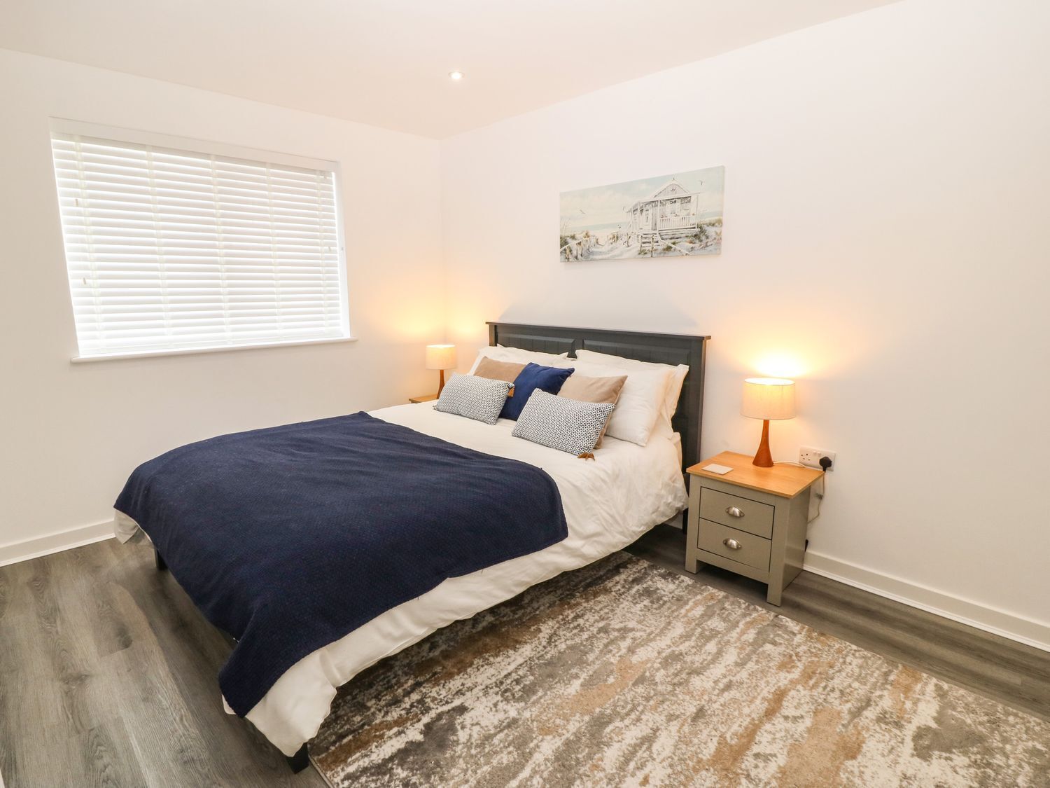 A bedroom with a bed, bedside lamps, a window with blinds, a painting on the wall, and a patterned rug.