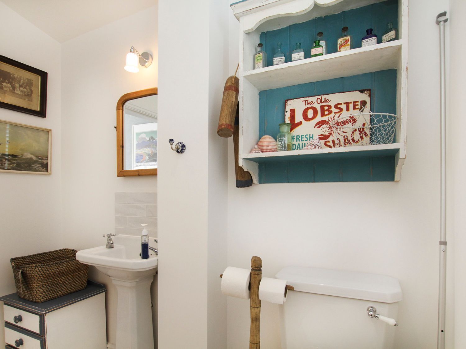 Un baño con lavamanos, espejo, cuadros enmarcados, repisas con botellas y un sujetapapeles.