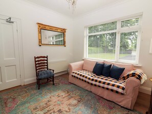 Ein Zimmer mit weißer Tür, einem Holzstuhl, einem rosa Sofa mit gemusterten Kissen, einem großen Spiegel und einem Fenster mit Jalousien.