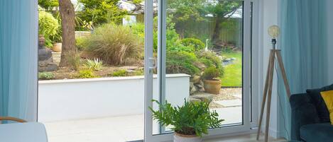 A room with a sliding glass door leading to a garden, a potted plant, a floor lamp, and a dark-colored sofa.