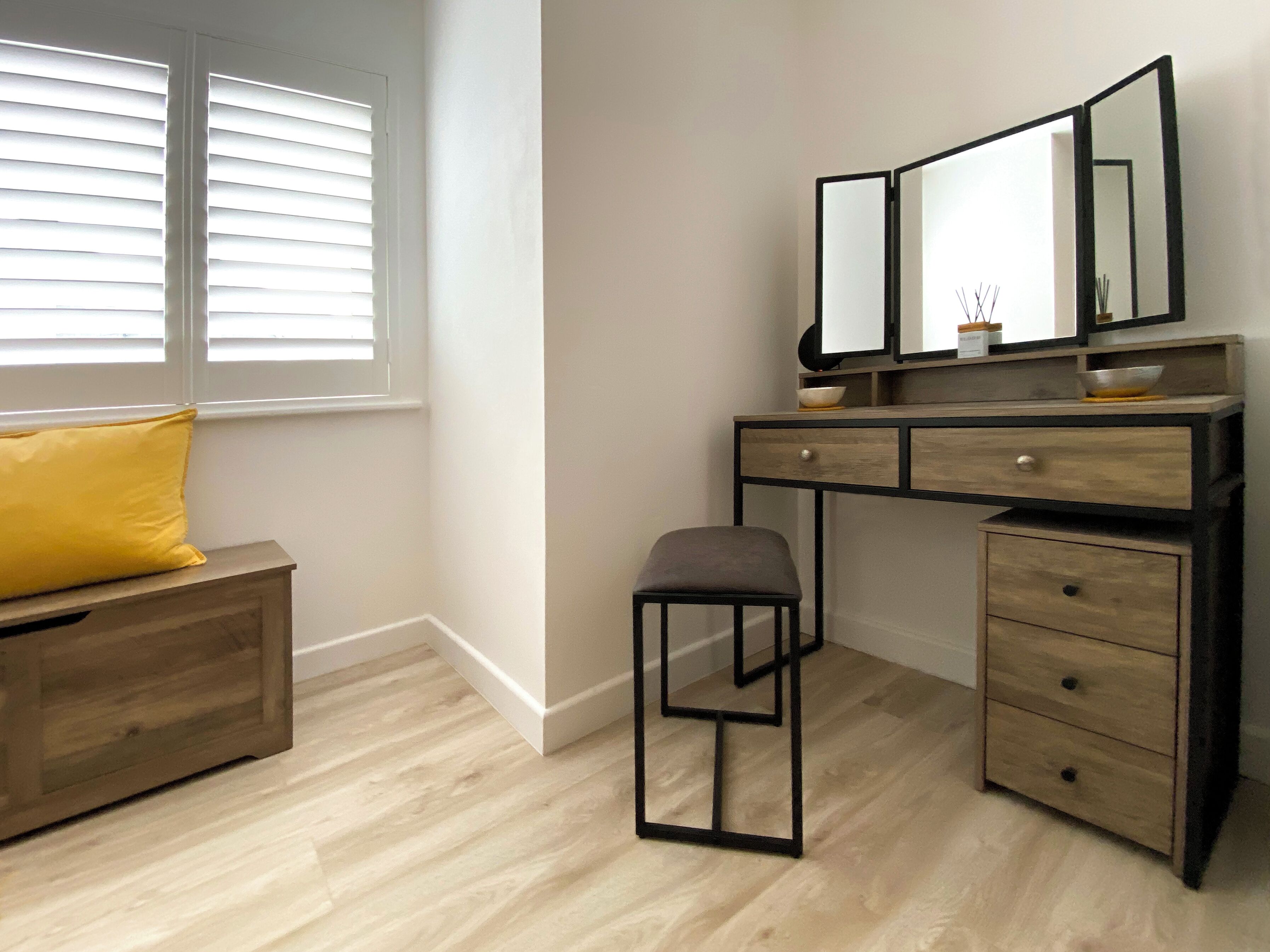 A bedroom with a wooden dresser, a small stool, a yellow pillow, and a window with blinds.