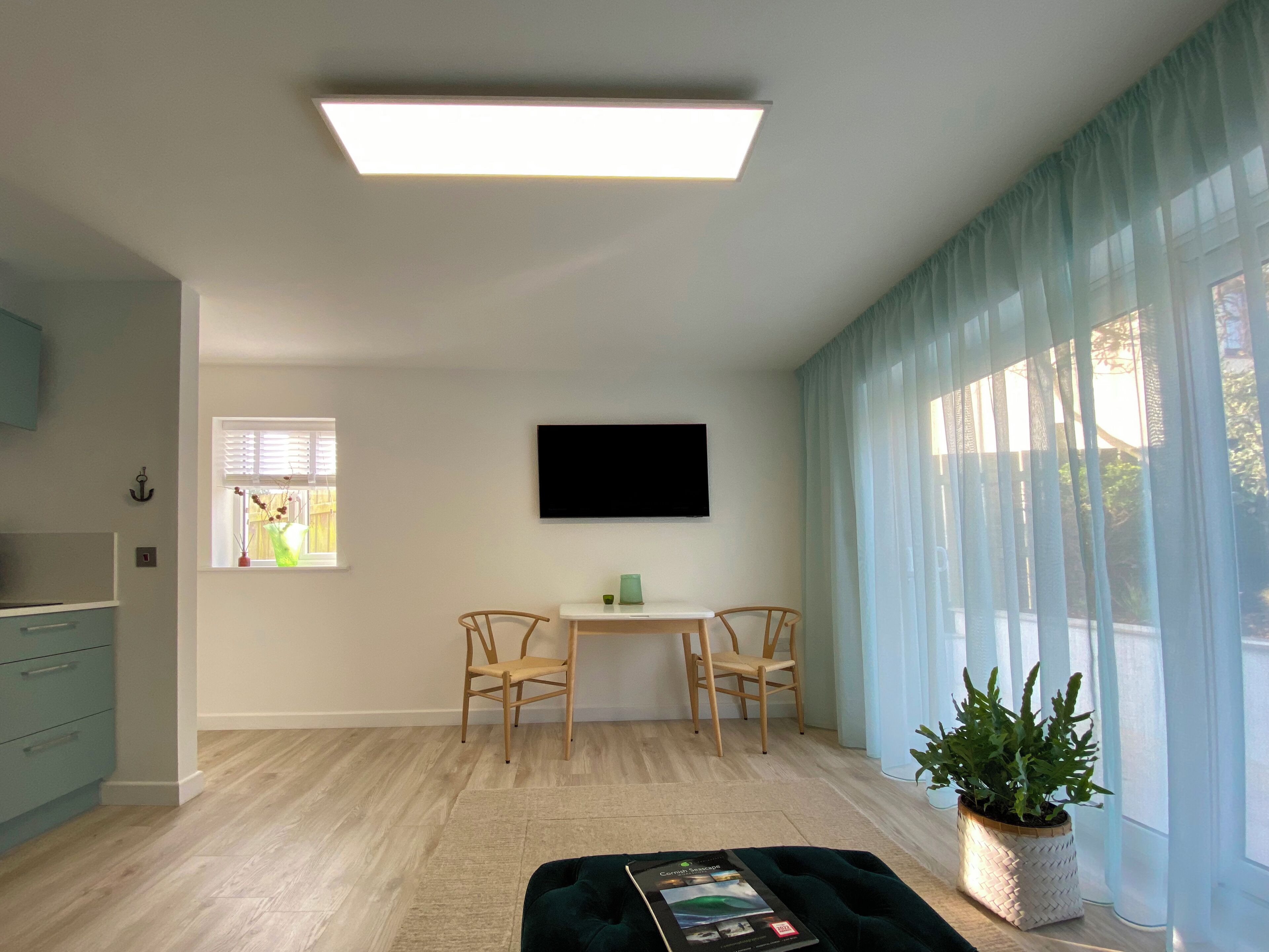A modern living room with a flat-screen TV mounted on the wall, two wooden chairs, a small table, a potted plant, and a sofa with a magazine.