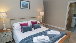 Una cama bien hecha con colcha azul, sábanas blancas y varias almohadas. Hay dos mesitas de noche con lámparas y un cuadro en la pared.