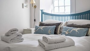 A neatly made bed with white and blue pillows, a lamp, and a window with a view. - Bodior Garden Cottage (Holyhead)