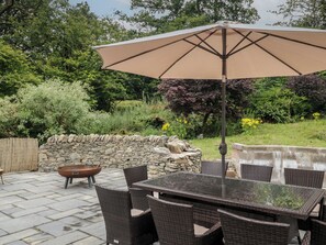 An outdoor patio with a table and chairs, a large umbrella, a stone wall, and a garden with various plants and trees.
